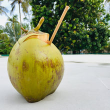 Load image into Gallery viewer, A bamboo straw is inserted into a cut coconut, with a background of greenery.